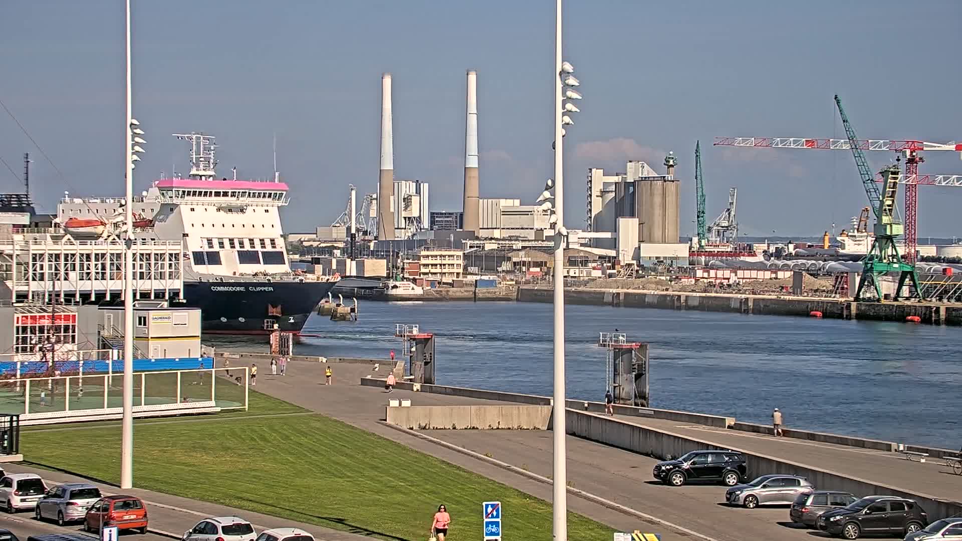 A ferry is docked at a port on a sunny day, with industrial buildings and cranes visible in the background.
