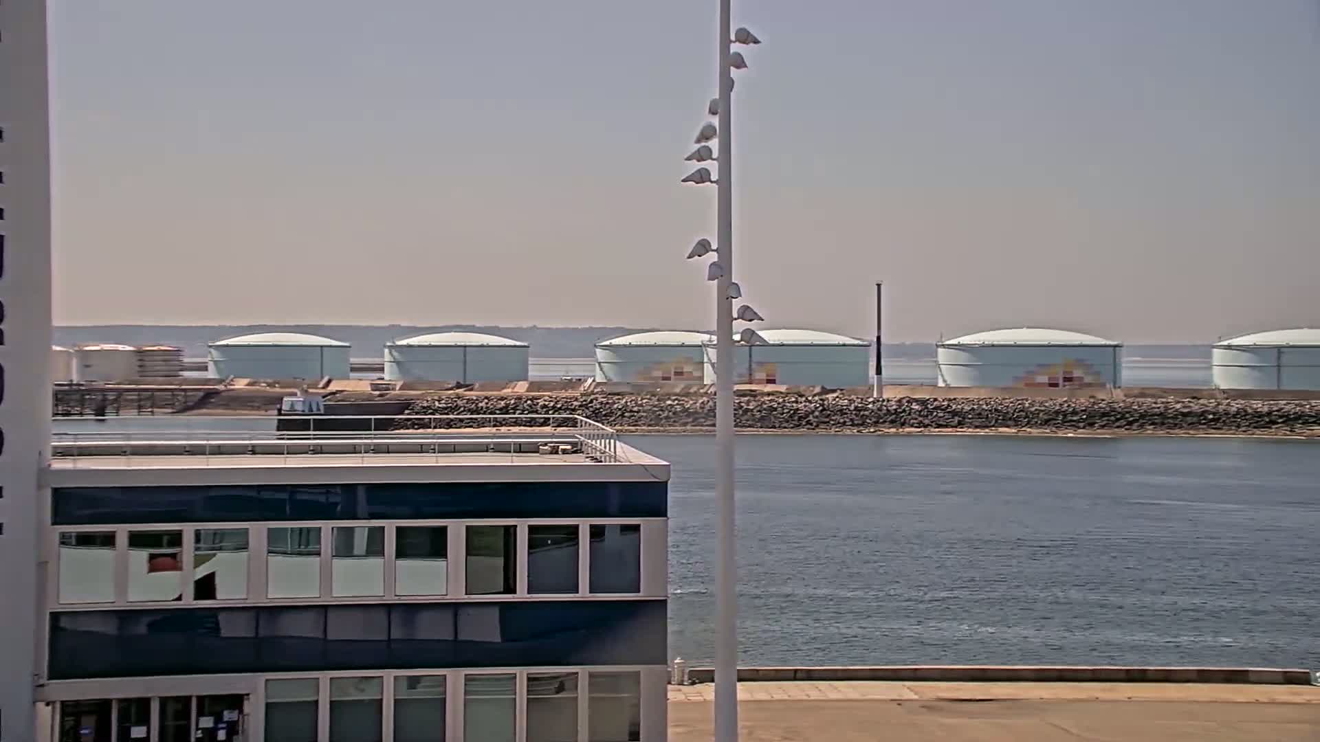 A row of large, light-blue storage tanks sits across a body of calm water from a modern building on a sunny day.