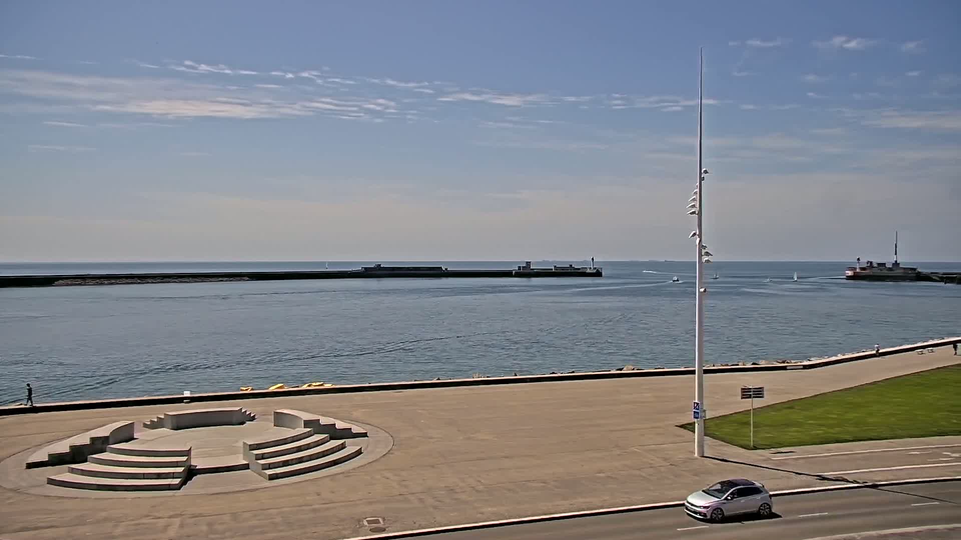 A harbor with calm water under a mostly sunny sky is viewed from a high vantage point, showing a paved area with a circular stone structure and a car driving along a road.
