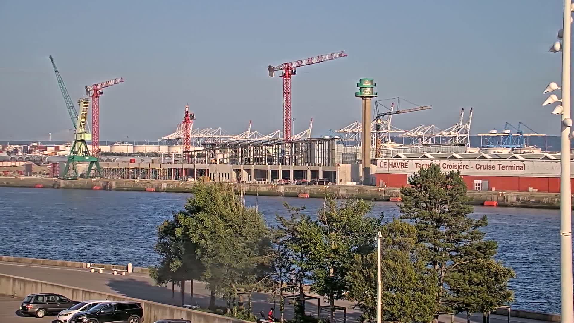 A waterfront construction site with several cranes and a partially built structure is visible across a calm body of water under a clear sky.