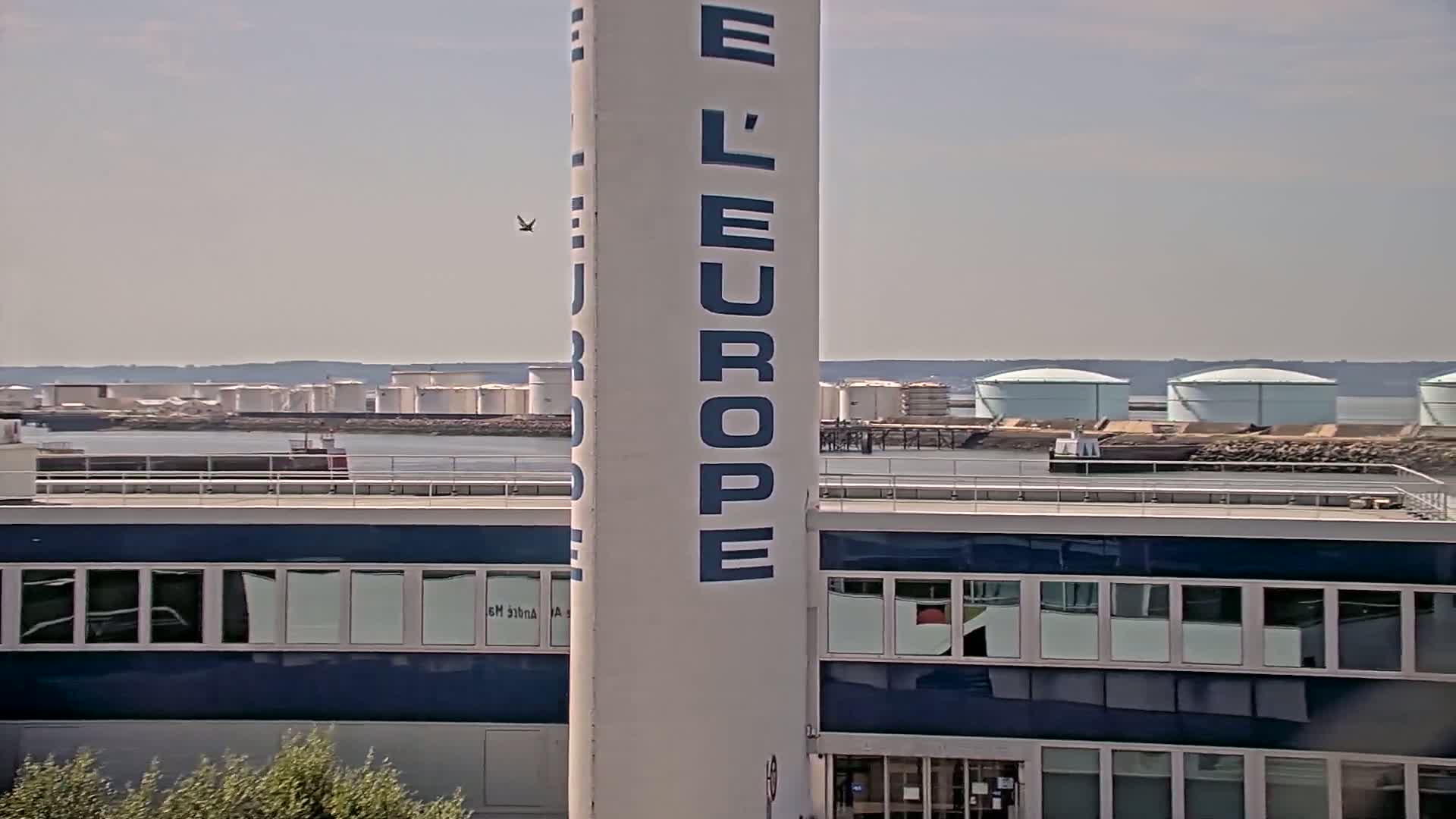 A tall, white tower with blue lettering stands in front of a blue building, overlooking a body of water with several large, white storage tanks in the distance under a clear sky.