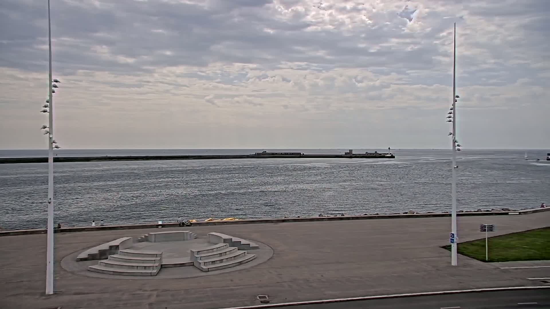 A stone amphitheater sits on a paved waterfront overlooking a calm sea under a cloudy sky.