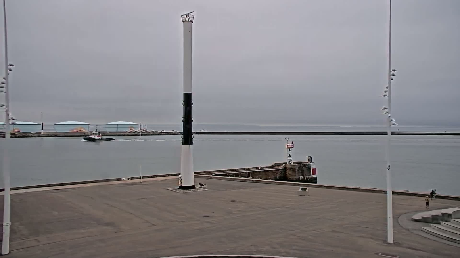 A tall, slender lighthouse with a black band stands in a harbor area on an overcast day, with a tugboat visible in the distance.