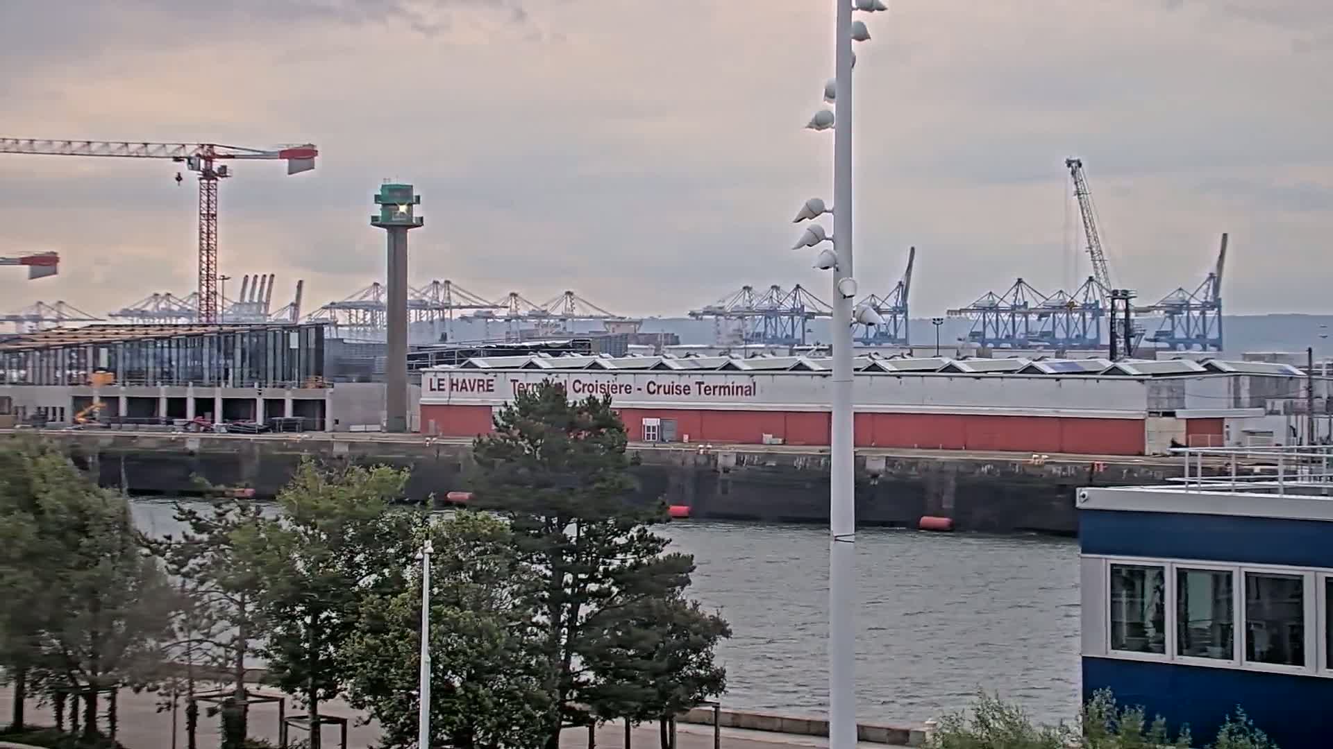 A cruise terminal sits on a waterfront, with numerous cranes and construction visible in the background under an overcast sky.