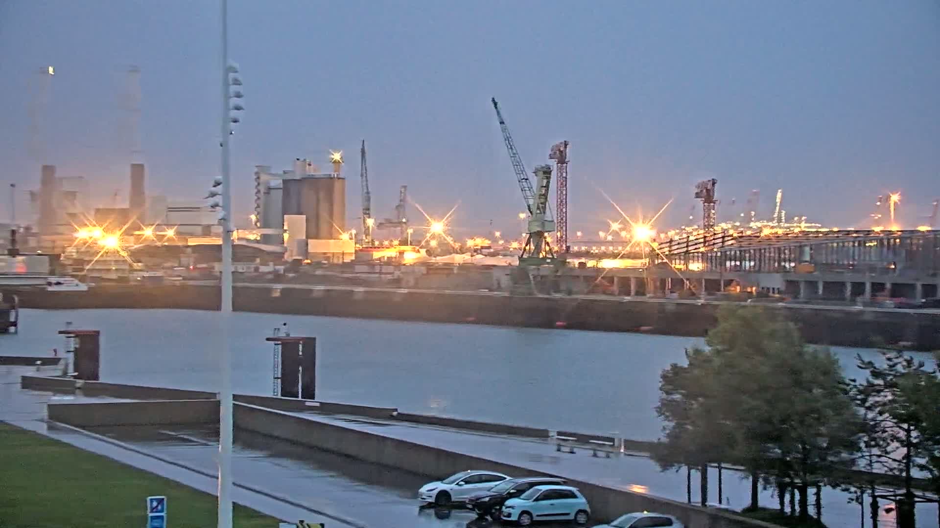 A rainy twilight scene shows an industrial port with cranes and buildings illuminated, a waterway in the foreground, and a few cars parked along a wet concrete area.