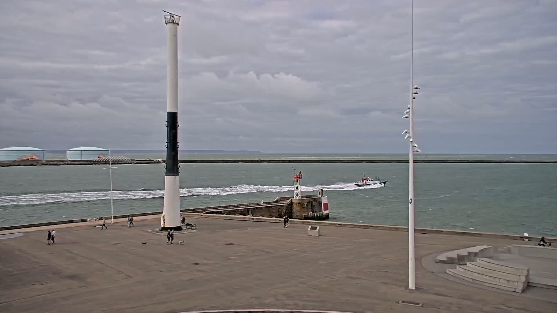 A tall, slender lighthouse stands on a paved waterfront area with a few pedestrians, overlooking a body of water where a small boat is moving, all under a cloudy sky.