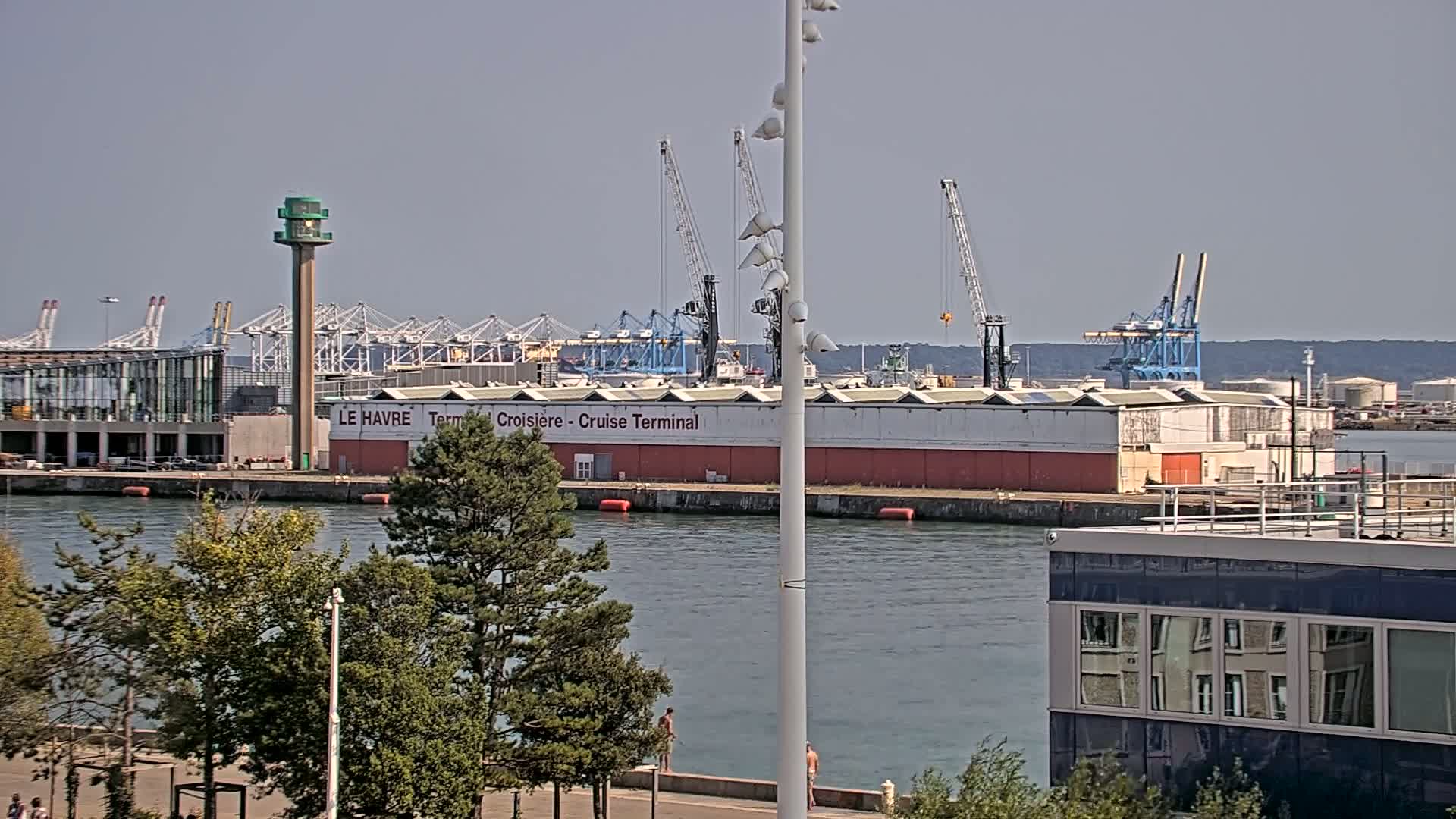 A tall, slender lighthouse stands on a paved waterfront area with a few pedestrians, overlooking a body of water where a small boat is moving, all under a cloudy sky.