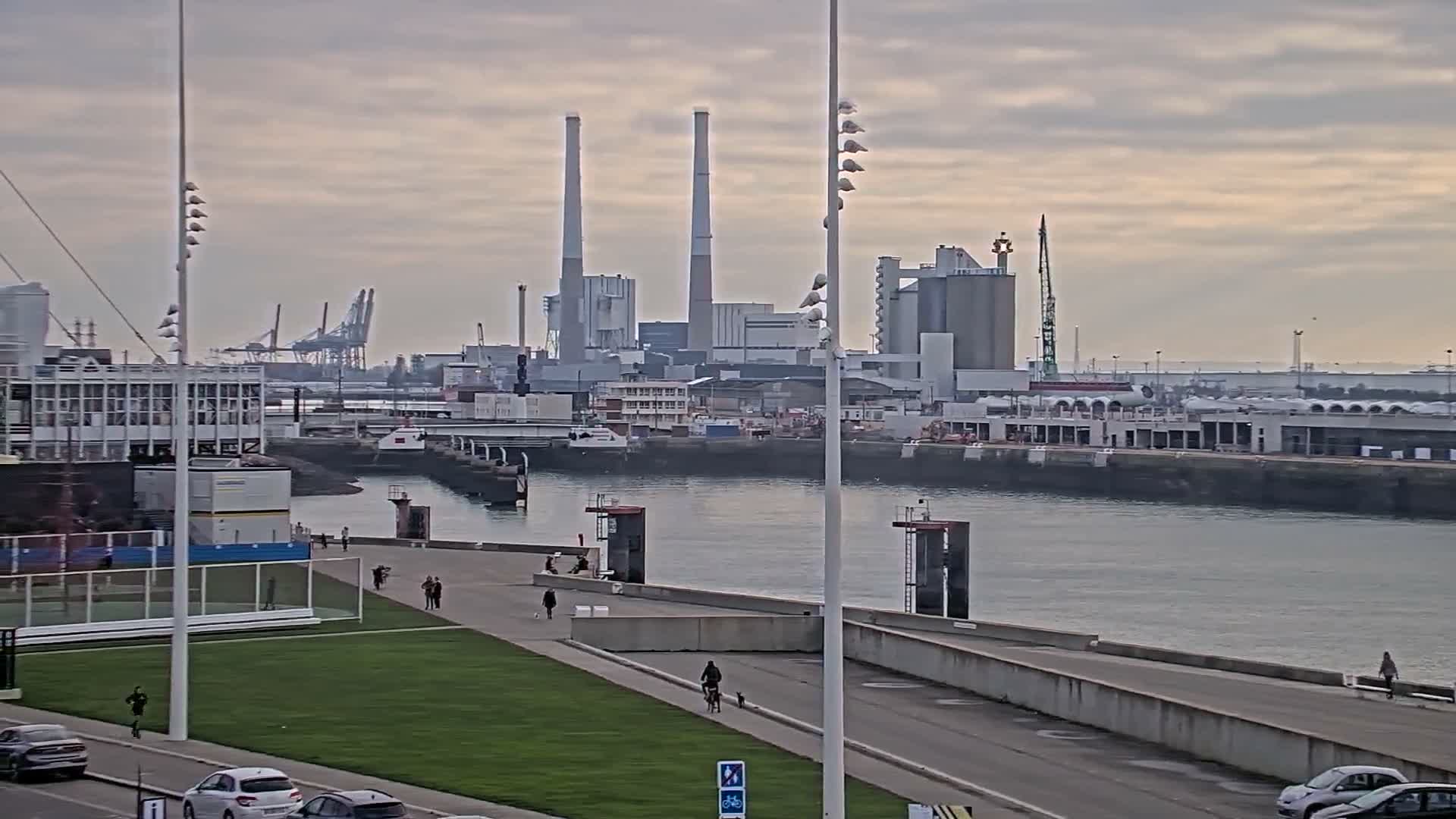 A tall, slender lighthouse stands on a paved waterfront area with a few pedestrians, overlooking a body of water where a small boat is moving, all under a cloudy sky.