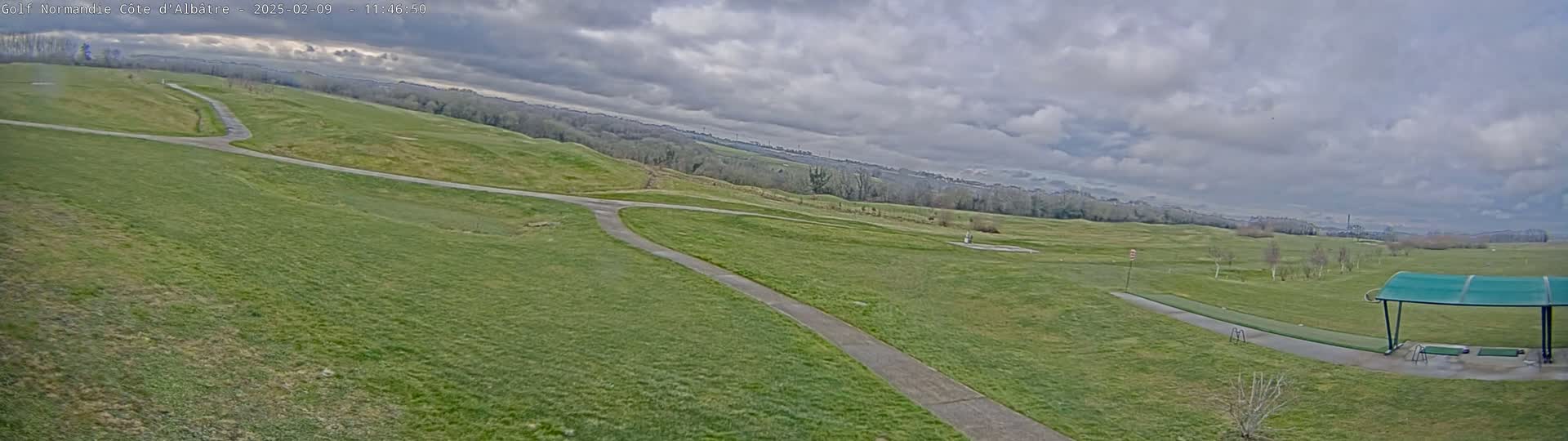 A mostly cloudy day reveals a view of a sprawling green golf course with winding pathways and a covered driving range in the foreground.