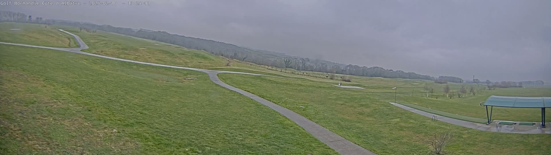 A wide, overcast view of a golf course with paved pathways and a covered driving range in the foreground.