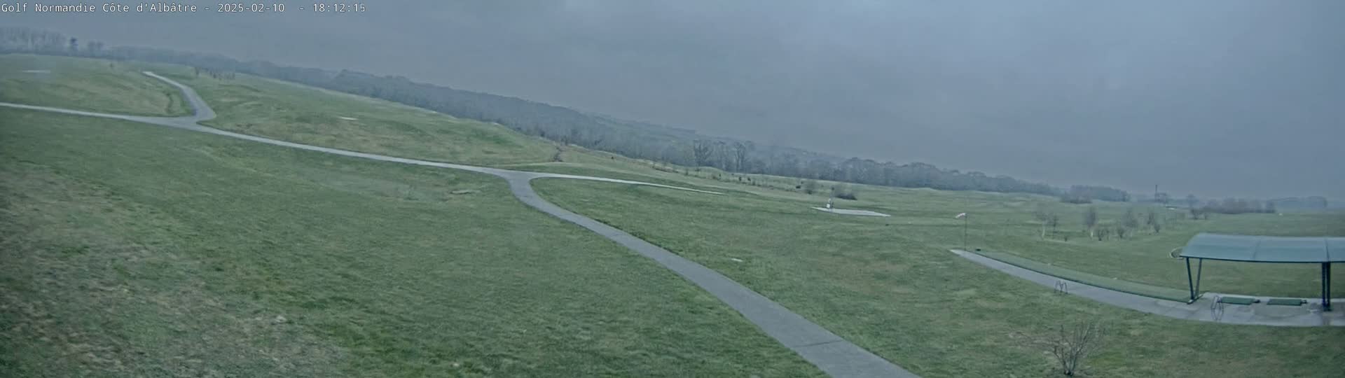 A mostly overcast view of a golf course with paved pathways and a covered driving range in the foreground.
