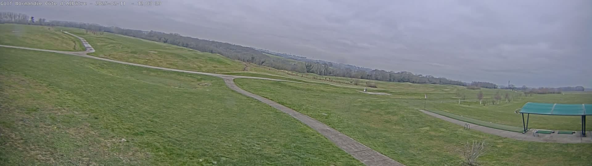 A sprawling green golf course stretches out under an overcast sky, featuring a paved pathway winding through the fairways and a covered driving range in the foreground.