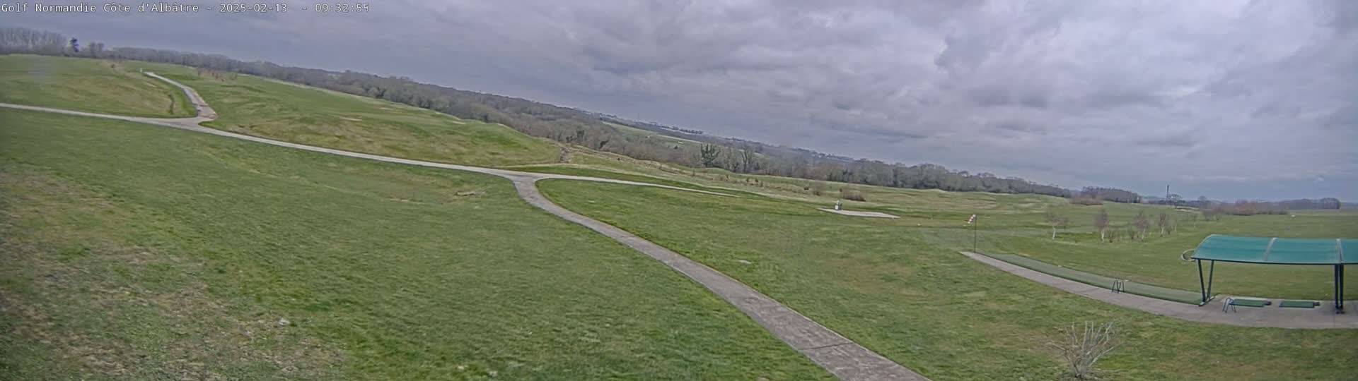 A view of a golf course on an overcast day shows two paved paths intersecting, leading to a driving range with a covered area.
