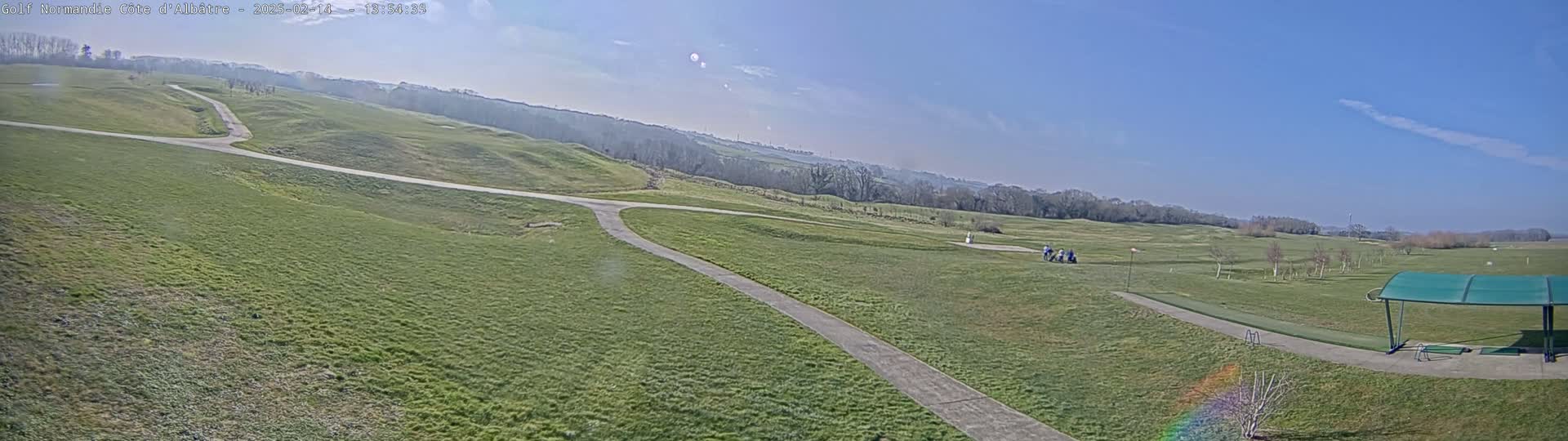A panoramic view of a golf course on a sunny day shows a wide expanse of green grass, winding cart paths, and a few people near a driving range.