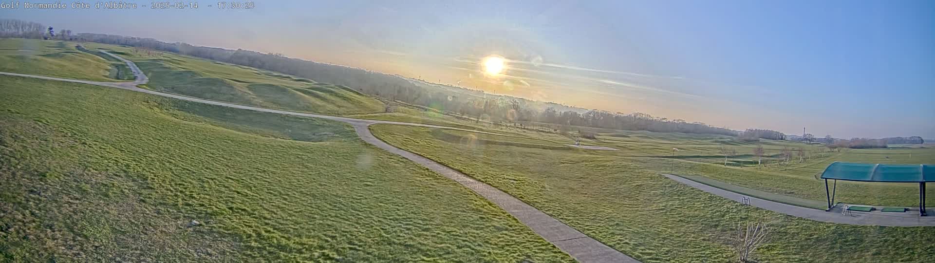 A sunny morning view of a golf course with rolling green hills, pathways, and a covered driving range.