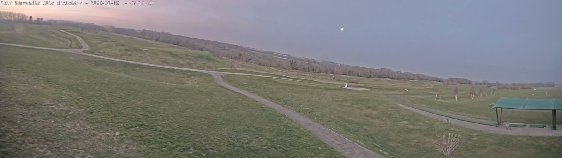 A mostly overcast sky hangs over a sprawling, hilly golf course with a paved path winding through it, culminating in a covered driving range.