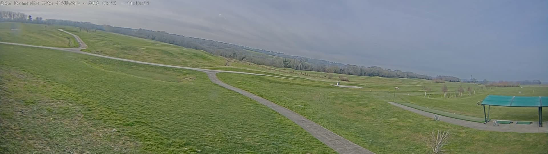 A mostly sunny day reveals a sprawling green golf course with paved pathways, a driving range, and a covered hitting area in the foreground.