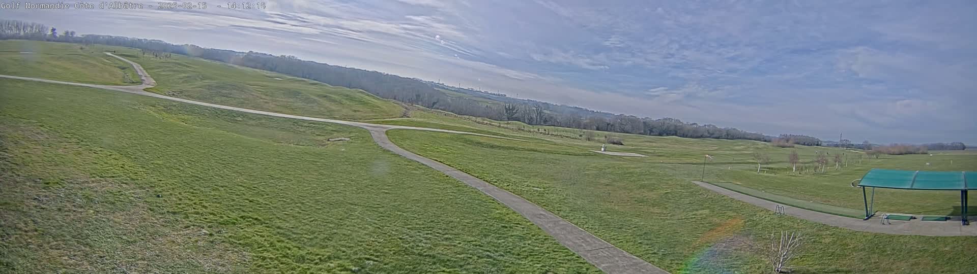 A mostly sunny day reveals a vast, green golf course with winding paths and a covered driving range in the foreground.