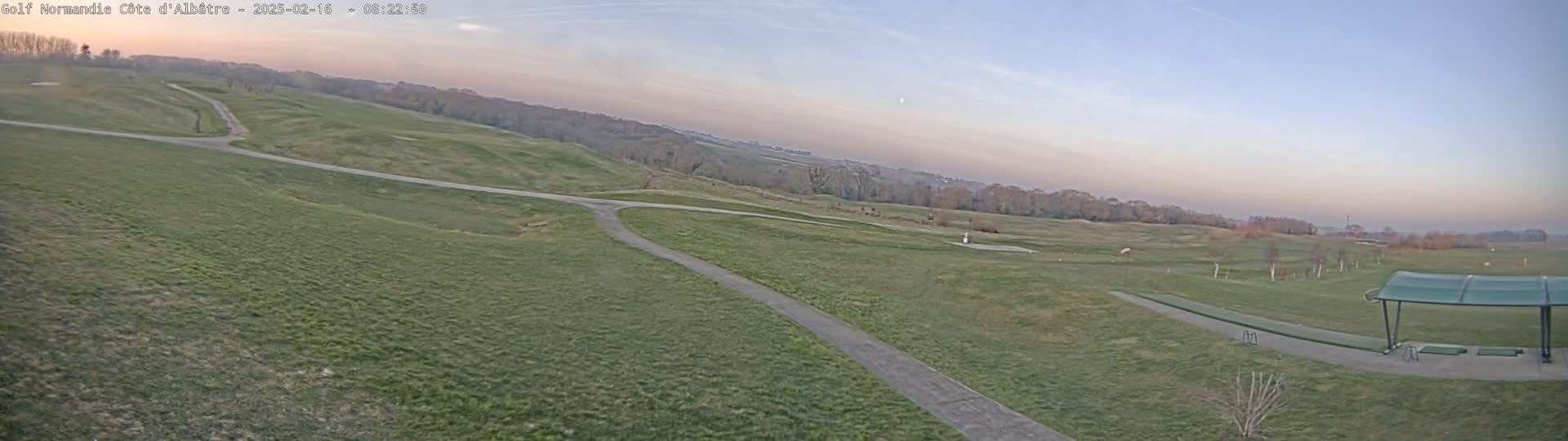 A wide, grassy expanse of a golf course, with paved paths and a covered driving range, under a clear, pale sky at dawn or dusk.