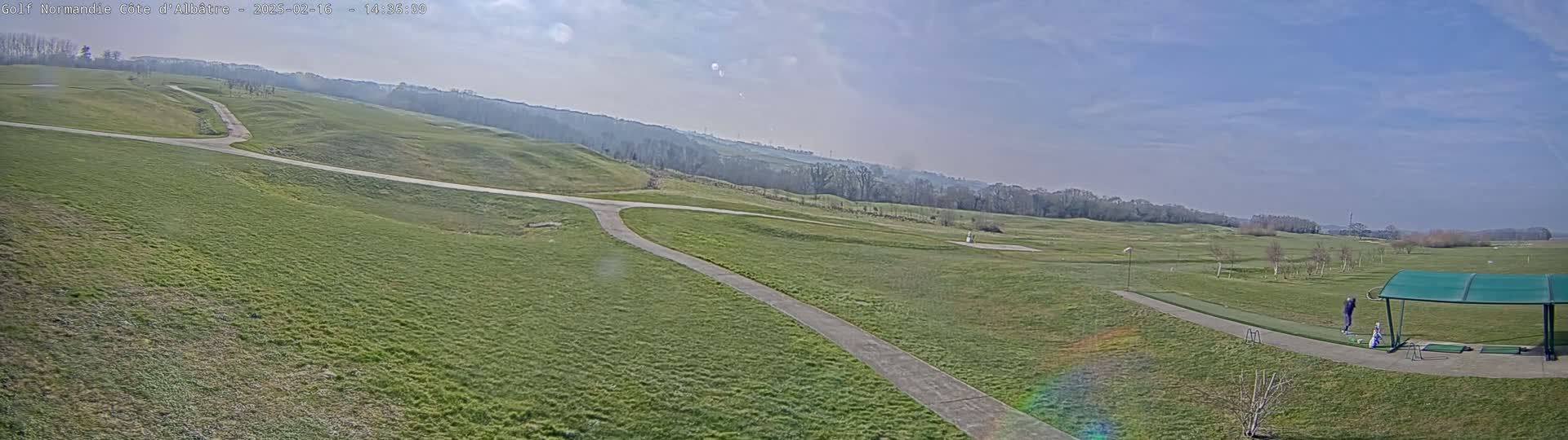 A mostly sunny day reveals a sprawling green golf course with paved pathways, a driving range with a covered area, and a few people practicing their golf swings.