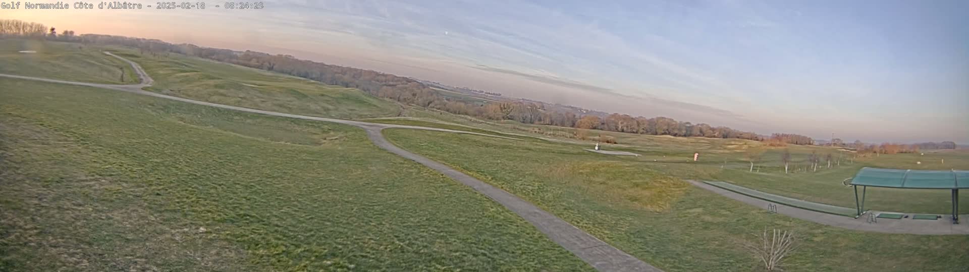 A panoramic view of a golf course under a partly cloudy, twilight sky shows a driving range with a covered hitting area and pathways winding through grassy fairways.