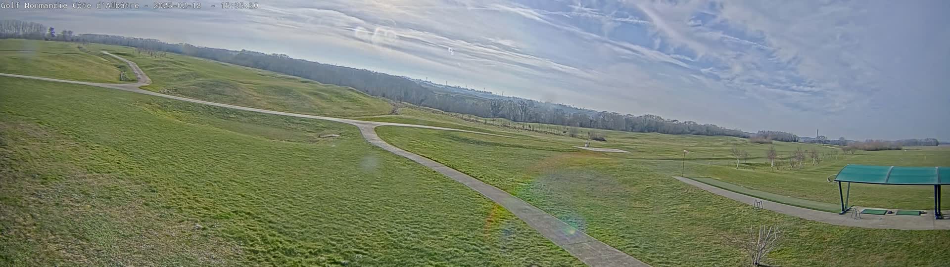 A mostly sunny day reveals a sprawling golf course with rolling green hills, paved pathways, and a covered driving range.
