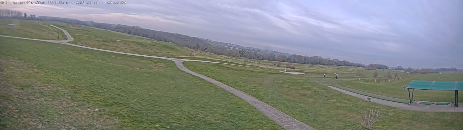 A golf course with a paved path and a covered driving range is visible under an overcast sky.