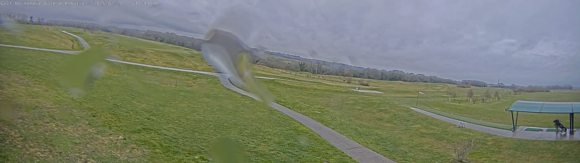 A view of a golf course on an overcast day, showing a path winding through green fairways and a driving range with a covered area.