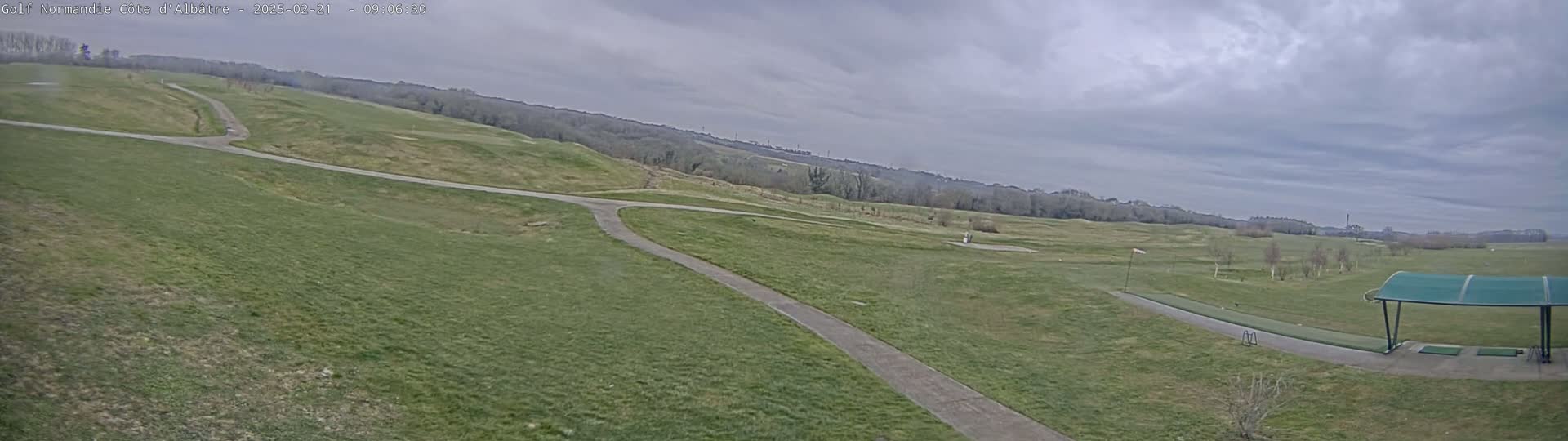 A wide shot of a golf course on an overcast day shows winding paths and greens, with a covered driving range in the lower right.