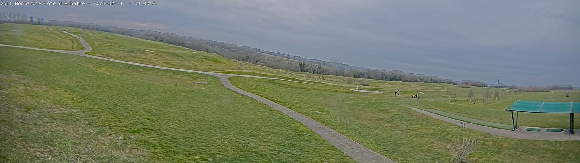 A wide, outdoor shot of a golf course on an overcast day shows a paved path winding through a grassy area, with golfers visible in the distance and a covered driving range in the lower right corner.