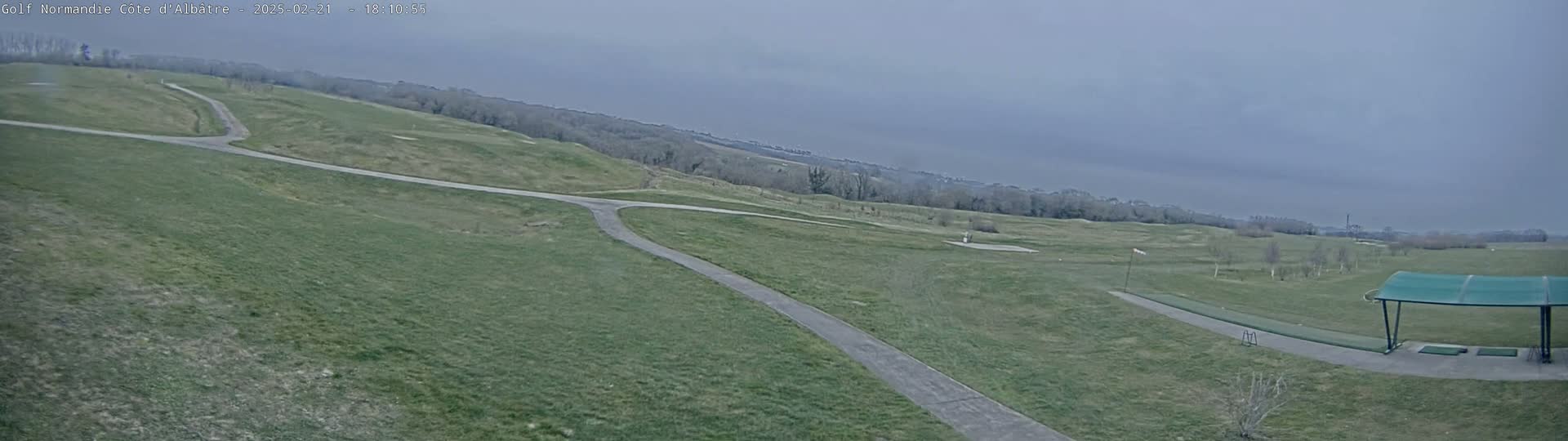 A mostly overcast sky hangs over a golf course with a paved path winding through grassy fairways and a covered driving range.