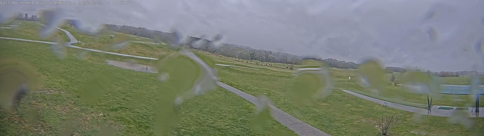 A rain-covered view of a golf course with pathways and a covered driving range in the distance under an overcast sky.