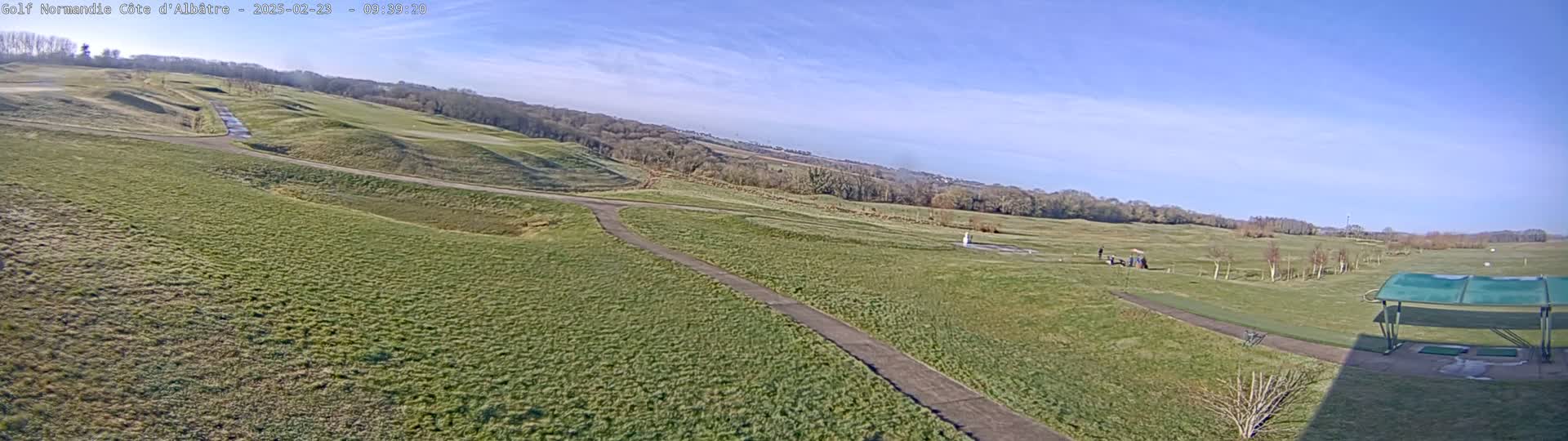 A sunny day reveals a sprawling golf course with rolling green hills, paved pathways, and a covered driving range in the foreground.