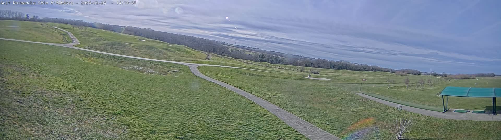 A mostly sunny day reveals a golf course with rolling green hills, paved pathways, and a covered driving range.