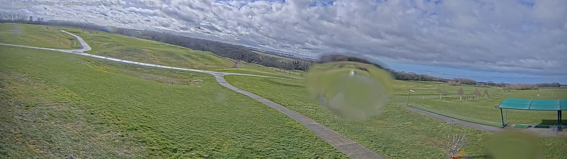 A mostly sunny day reveals a panoramic view of a golf course with rolling green hills, a paved path, and a covered driving range.