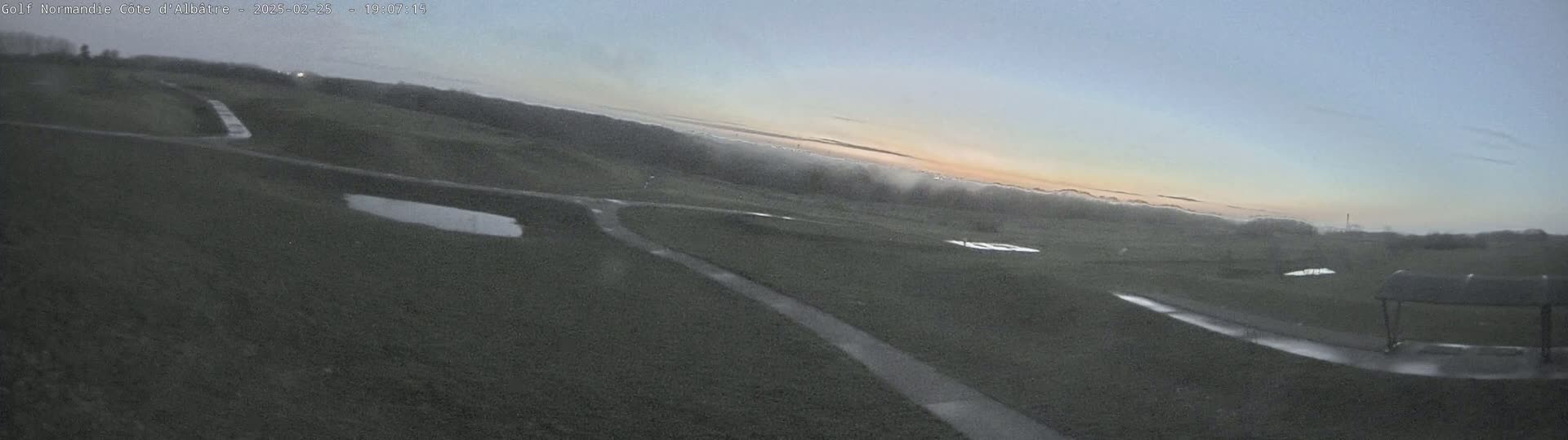 A mostly overcast sky with a hint of sunset illuminates a dark green golf course with paved pathways and small puddles of water.