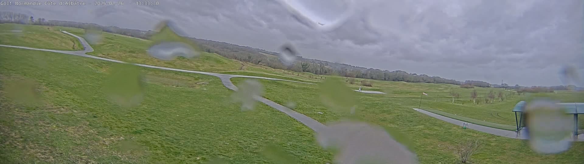 A rain-covered view of a golf course with paved pathways winding through grassy hills under a cloudy sky.