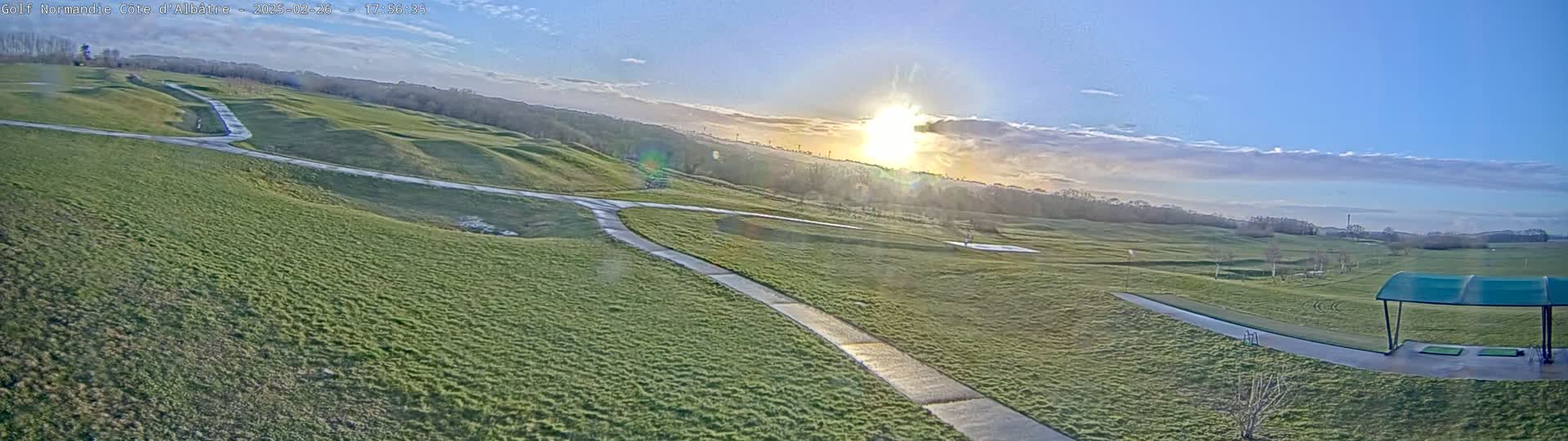 A sunny, partly cloudy day reveals a golf course with rolling green hills, paths, and a covered driving range.