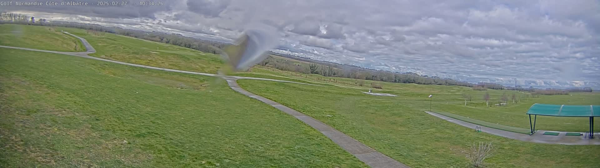 A view of a golf course on an overcast day shows a paved path winding through a green grassy area, with a driving range visible in the distance under a covered shelter.