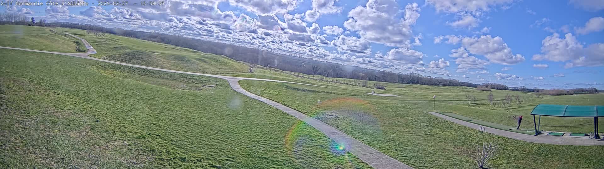 A partly sunny day at a golf course shows rolling green hills, paved paths, and a person practicing at a covered driving range.