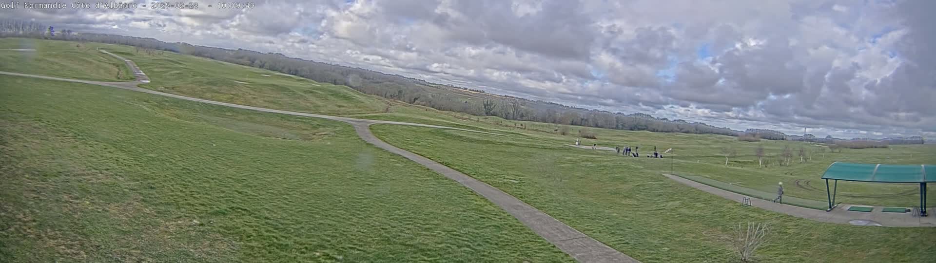 A sprawling green golf course with paved pathways, under a cloudy sky, features several people near a driving range.