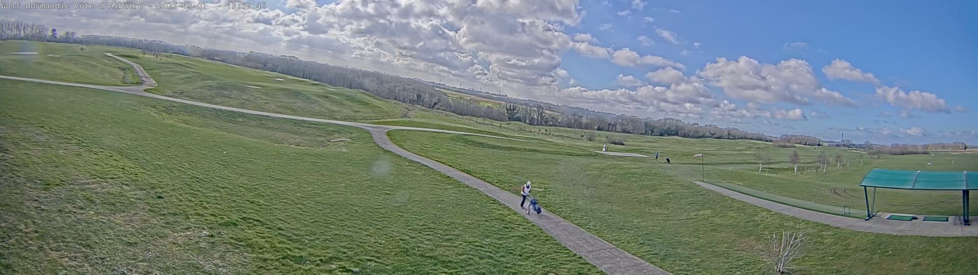 A person pulls a golf cart along a paved path on a partly cloudy day across a sprawling green golf course.