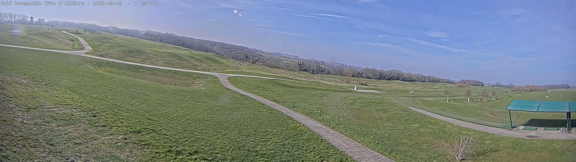 A mostly sunny day reveals a sprawling green golf course with winding paths and a covered driving range in the foreground.