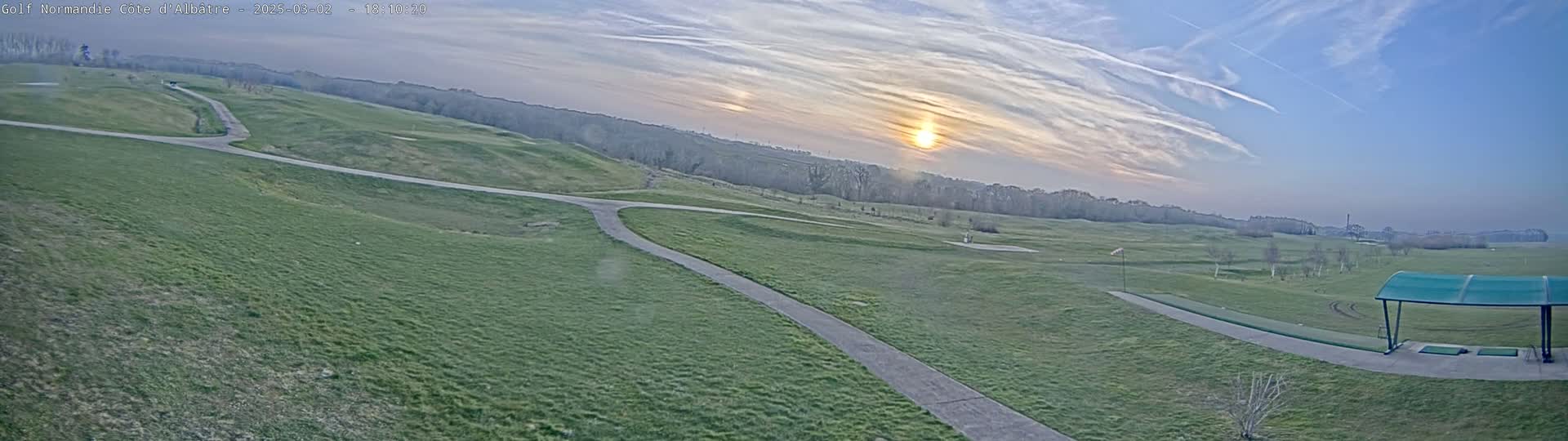 A wide, expansive view of a golf course under a partly cloudy sky, with the sun low on the horizon.
