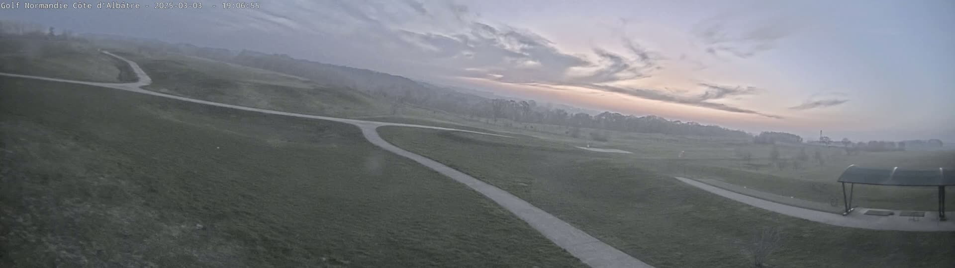 A hazy sunrise illuminates a golf course with winding paths and a covered structure in the distance.