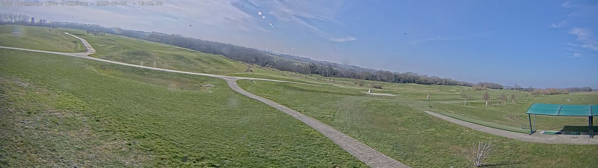 A mostly sunny day reveals a sprawling golf course with paved pathways winding through grassy fairways, a driving range with a covered shelter, and a distant tree line.
