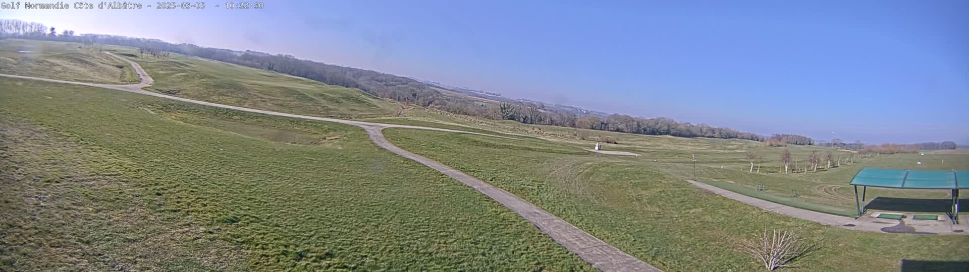 A panoramic view of a golf course on a sunny day shows rolling green fairways, paved paths, and a covered driving range.