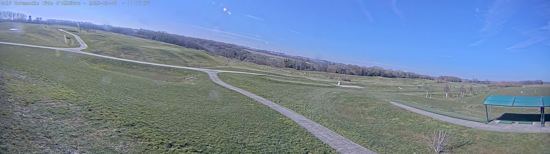 A mostly sunny, outdoor view of a golf course with a driving range and a covered area for golfers, showing paths winding through the grassy terrain.