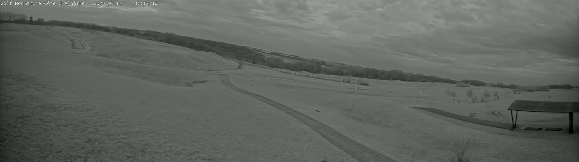 A mostly cloudy sky hangs over a hilly, snow-covered golf course with a paved path and a covered structure in the foreground.