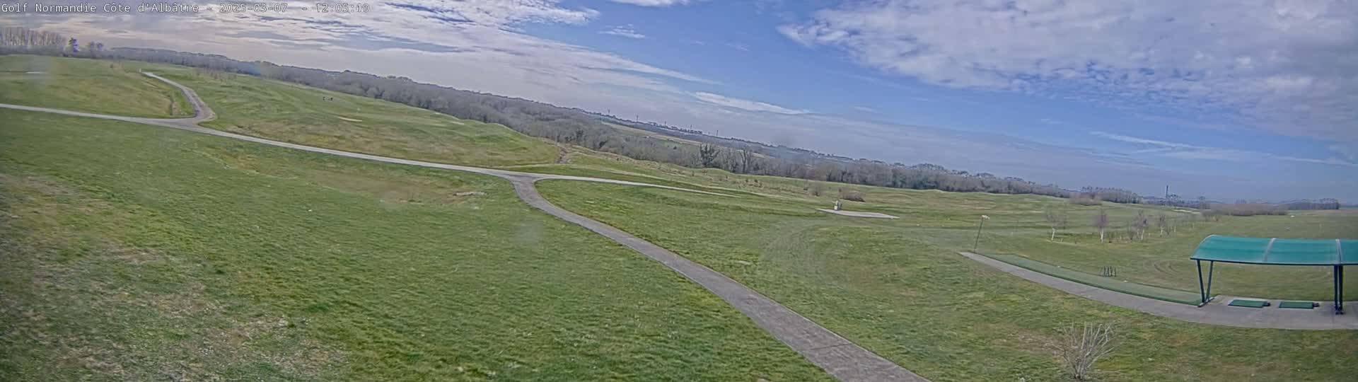 A partly cloudy sky overlooks a sprawling green golf course with paved pathways, and a covered driving range in the foreground.